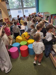Na zdjęciu znajdują się uczestnicy zajęć. Dzieci wykonują zadania pomiędzy regałami z książkami.  Zdjęcie wykonano w bibliotece.