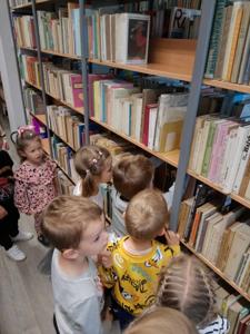Na zdjęciu znajdują się uczestnicy zajęć. Dzieci wykonują zadania pomiędzy regałami z książkami.  Zdjęcie wykonano w bibliotece.