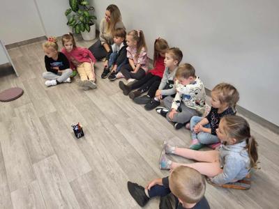 Na zdjęciu znajdują się uczestnicy zajęć. Dzieci wykonują zadania siedząc na poduszkach. Zdjęcie wykonano w bibliotece.