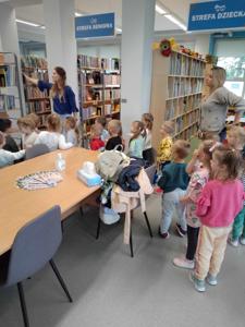Na zdjęciu znajdują się uczestnicy zajęć. Dzieci wykonują zadania pomiędzy regałami z książkami.  Zdjęcie wykonano w bibliotece.