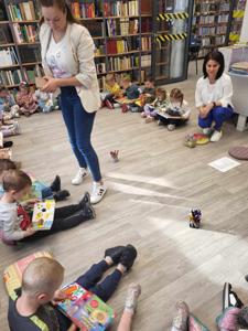 Na zdjęciu znajdują się uczestnicy zajęć. Dzieci wykonują zadania siedząc na poduszkach. Zdjęcie wykonano w bibliotece.