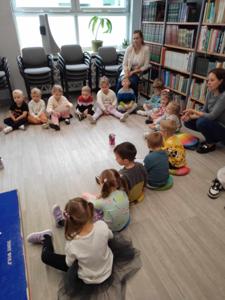 Na zdjęciu znajdują się uczestnicy zajęć. Dzieci wykonują zadania siedząc na poduszkach. Zdjęcie wykonano w bibliotece.