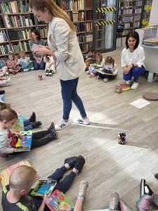Na zdjęciu znajdują się uczestnicy zajęć. Dzieci wykonują zadania siedząc na poduszkach. Zdjęcie wykonano w bibliotece.