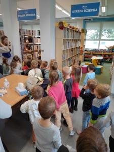 Na zdjęciu znajdują się uczestnicy zajęć. Dzieci wykonują zadania pomiędzy regałami z książkami.  Zdjęcie wykonano w bibliotece.