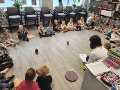 Na zdjęciu znajdują się uczestnicy zajęć. Dzieci wykonują zadania siedząc na poduszkach. Zdjęcie wykonano w bibliotece.
