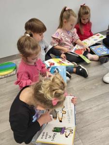 Na zdjęciu znajdują się uczestnicy zajęć. Dzieci wykonują zadania siedząc na poduszkach. Zdjęcie wykonano w bibliotece.