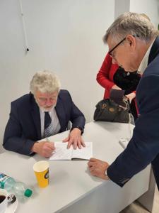 Na zdjęciu znajduje się autor Zbigniew Kołba trzymający książkę. W tle uczestnicy spotkania.