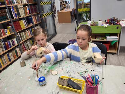 Na zdjęciu znajdują się uczestnicy warsztatów. Młodzież siedzi i wykonuje rękodzieło. Zdjęcie wykonano w bibliotece.