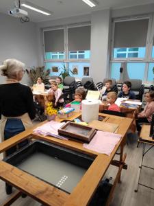 Na zdjęciu znajdują się uczestnicy warsztatów. Dzieci siedzą przy stole i piszą. Zdjęcie wykonano w bibliotece.