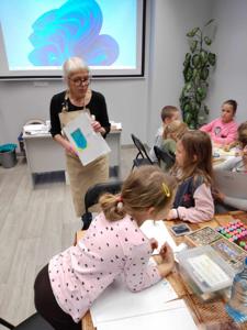 Na zdjęciu znajdują się uczestnicy warsztatów. Dzieci siedzą przy stole i piszą. Zdjęcie wykonano w bibliotece.