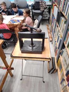 Na zdjęciu znajdują się uczestnicy warsztatów. Dzieci siedzą przy stole i piszą. Zdjęcie wykonano w bibliotece.
