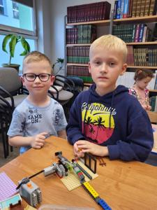 Na zdjęciu znajdują się uczestnicy warsztatów. Dzieci siedzą i stoją przy stole wykonując konstrukcje z klocków. Zdjęcie wykonano w bibliotece.