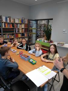 Na zdjęciu znajdują się uczestnicy warsztatów. Młodzież siedzi przy stole, wykonuje oraz ogląda komiksy. Zdjęcie wykonano w bibliotece.