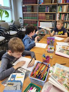 Na zdjęciu znajdują się uczestnicy warsztatów. Młodzież siedzi przy stole, wykonuje oraz ogląda komiksy. Zdjęcie wykonano w bibliotece.