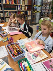 Na zdjęciu znajdują się uczestnicy warsztatów. Młodzież siedzi przy stole, wykonuje oraz ogląda komiksy. Zdjęcie wykonano w bibliotece.