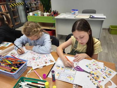 Na zdjęciu znajdują się uczestnicy warsztatów. Młodzież siedzi przy stole, wykonuje oraz ogląda komiksy. Zdjęcie wykonano w bibliotece.