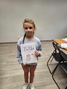 Na zdjęciu znajduje się uczestniczka warsztatów. Dziewczyna trzyma w ręce komiks. Zdjęcie wykonano w bibliotece.