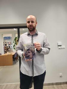 Na zdjęciu znajduje się prelegent. Prelegent trzyma w rękach książkę. W tle księgozbiór biblioteki.