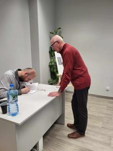 Na zdjęciu znajduje się prelegent siedzący przy biurku. Jedna osoba stoi przy biurku.