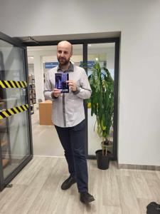 Na zdjęciu znajduje się prelegent. Prelegent trzyma w rękach książkę. W tle księgozbiór biblioteki.