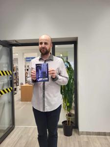 Na zdjęciu znajduje się prelegent. Prelegent trzyma w rękach książkę. W tle księgozbiór biblioteki.