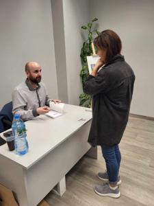 Na zdjęciu znajduje się prelegent siedzący przy biurku. Jedna osoba stoi przy biurku.