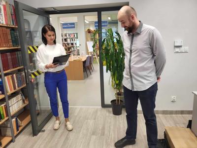 Ma zdjęciu znajdują się dwie osoby. W tle księgozbiór biblioteki.