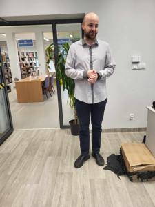 Na zdjęciu znajduje się prelegent. Prelegent trzyma w rękach książkę. W tle księgozbiór biblioteki.