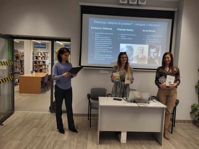 Na zdjęciu znajdują się prelegentki. Zdjęcie wykonano w bibliotece. W tle prezentacja multimedialna.