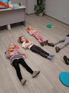 Na zdjęciu znajdują się uczestnicy warsztatów. Dzieci leżą na podłodze mając na sobie jabłko.