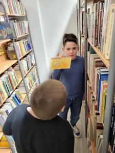 Na zdjęciu znajdują się uczestnicy warsztatów. Dzieci stoją. W tle księgozbiór biblioteki.