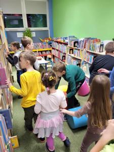 Na zdjęciu znajdują się uczestnicy zajęć. Dzieci stoją pomiędzy regałami.