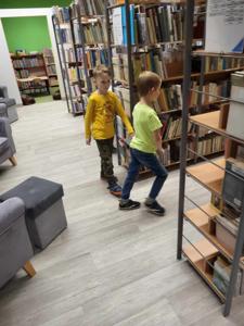 Na zdjęciu znajdują się uczestnicy warsztatów. Dzieci stoją. W tle księgozbiór biblioteki.