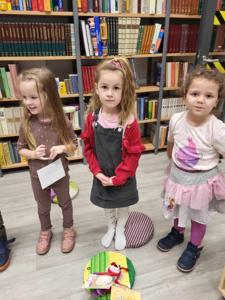 Na zdjęciu znajdują się uczestnicy warsztatów. Dzieci stoją. W tle księgozbiór biblioteki.