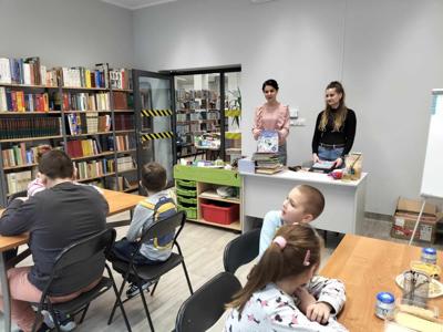 Na zdjęciu znajdują się uczestnicy zajęć. Dzieci siedzą przy stołach. Prowadzące stoją i pokazują książki uczestnikom.