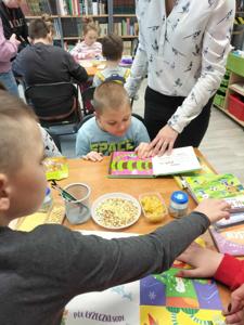 Na zdjęciu znajdują się uczestnicy zajęć. Dzieci siedzą przy stołach i oglądają książki.