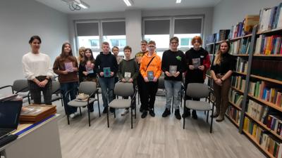 Na zdjęciu znajdują się uczestnicy wykładu. Uczestnicy stoją i trzymają książki w ręce. W tle księgozbiór biblioteki.