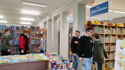 Na zdjęciu znajdują się uczestnicy wykładu. Młodzież znajduje się pomiędzy regałami. W tle księgozbiór biblioteki.