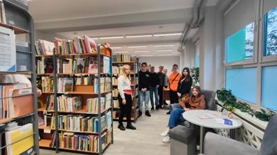 Na zdjęciu znajdują się uczestnicy wykładu. Młodzież znajduje się pomiędzy regałami. W tle księgozbiór biblioteki.