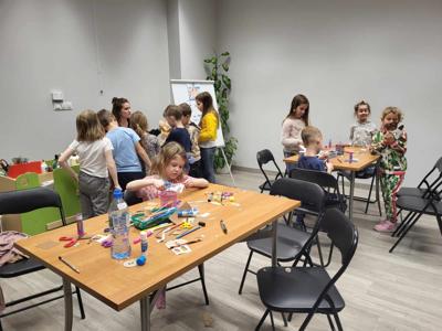 Na zdjęciu znajdują się uczestnicy warsztatów. Dzieci siedzą przy stole i wykonują prace plastyczne. Zdjęcie wykonano w bibliotece.