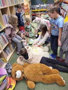 Na zdjęciu znajdują się uczestnicy warsztatów. Dzieci stoją i leżą z misiami. Zdjęcie wykonano w bibliotece.