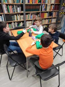 Na zdjęciu znajdują się uczestnicy warsztatów. Dzieci wykonują prace plastyczne siedząc przy stole. Zdjęcie wykonano w bibliotece.