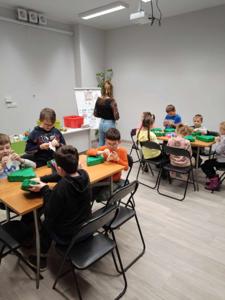 Na zdjęciu znajdują się uczestnicy warsztatów. Dzieci wykonują prace plastyczne siedząc przy stole. Zdjęcie wykonano w bibliotece.
