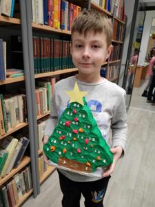 Na zdjęciu znajdują się uczestnicy warsztatów. Dzieci trzymają w rękach choinki. W tle księgozbiór biblioteki. Zdjęcie wykonano w bibliotece.