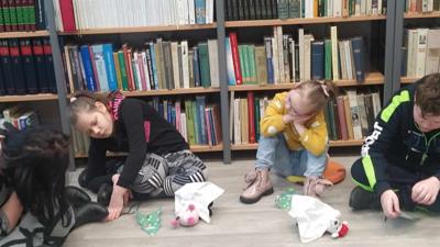 Na zdjęciu znajdują się uczestnicy zajęć. Dzieci siedzą na podłodze. Zdjęcie wykonano w bibliotece. W tle księgozbiór biblioteki.