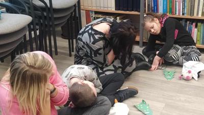 Na zdjęciu znajdują się uczestnicy zajęć. Dzieci siedzą na podłodze. Zdjęcie wykonano w bibliotece. W tle księgozbiór biblioteki.