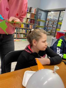 Na zdjęciu znajdują się uczestnicy zajęć. Dzieci siedzą przy stole. Zdjęcie wykonano w bibliotece. W tle księgozbiór biblioteki.