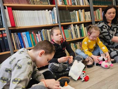 Na zdjęciu znajdują się uczestnicy zajęć. Dzieci siedzą na podłodze. Zdjęcie wykonano w bibliotece. W tle księgozbiór biblioteki.