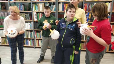 Na zdjęciu znajdują się uczestnicy zajęć. Dzieci stoją. Zdjęcie wykonano w bibliotece. W tle księgozbiór biblioteki.