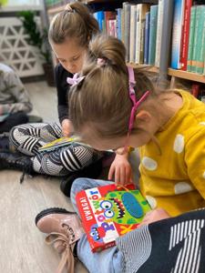 Na zdjęciu znajdują się uczestnicy zajęć. Dzieci siedzą na podłodze. Zdjęcie wykonano w bibliotece. W tle księgozbiór biblioteki.